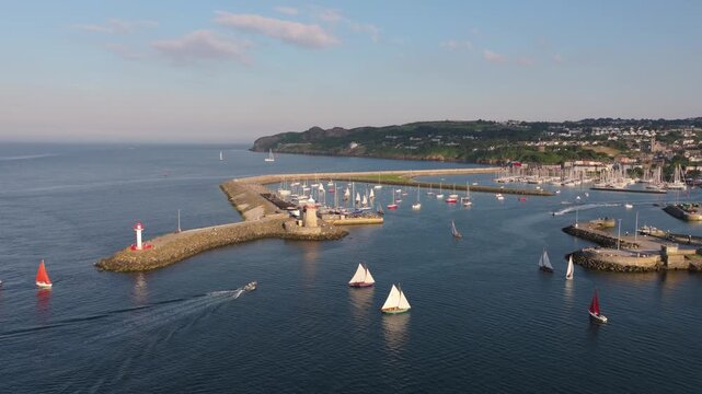 Small sailboats and a motorboat move past twin breakwater lighthouses at Howth Harbour as the drone glides over the calm evening water.