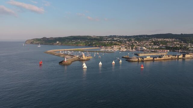 Small sailing boats and a motorboat move through the Howth harbour entrance beside the pier lighthouses on a clear day.