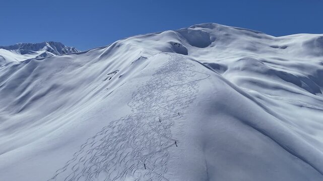 Aerial drone footage follows a group of ski tourers ascending a snow-covered ridge toward the summit, with numerous ski tracks visible across the surrounding alpine slopes near Ushguli in the Caucasus