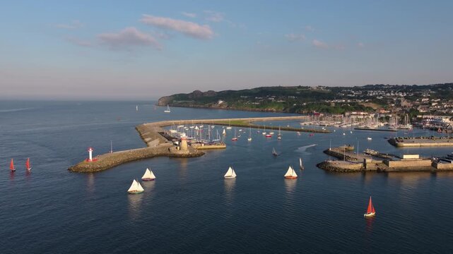 Colorful sailboats cruise into Howth Harbour past twin lighthouse breakwaters under warm evening light.