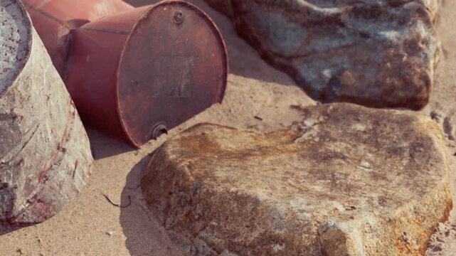 Barrels rest on the sand beside rocks at a beach. The sun shines down, creating a warm atmosphere. The scene shows signs of weathering from sun and sand.