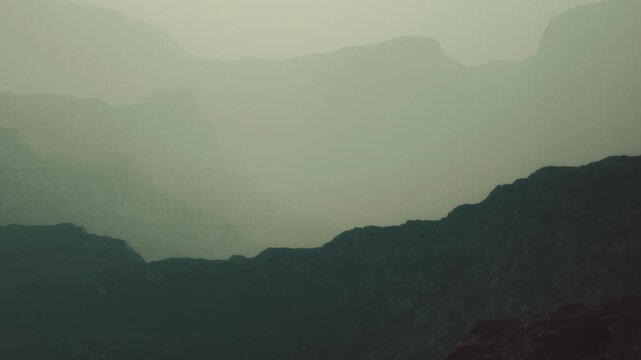 Fog spreads across mountains as the sun begins to rise. The landscape shows different layers of hills and valleys. The scene has limited visibility due to the fog.