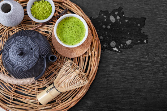 Matcha tea served in white ceramic bowl with bamboo whisk on wicker tray and nearby there is a puddle of spilled tea. Cast iron teapot and powdered green tea leaves for traditional