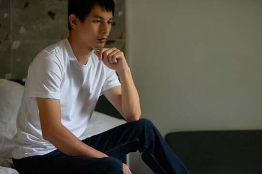 Thoughtful young man in casual clothing sitting on bed indoors