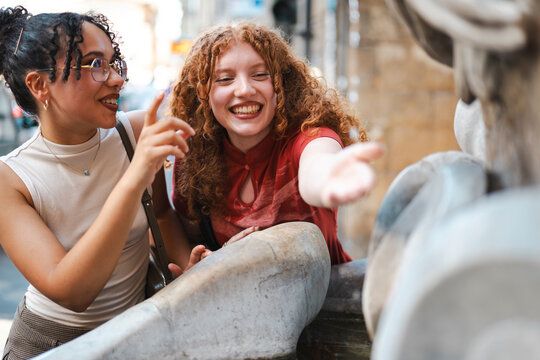 Two happy diverse female friends enjoy sightseeing together in a historic European city, laughing as they admire and interact with an old stone fountain.