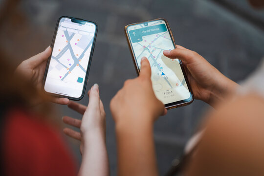 Two female travelers hold up their smartphones side by side to compare digital map navigation routes while searching for directions on a cobblestone street.