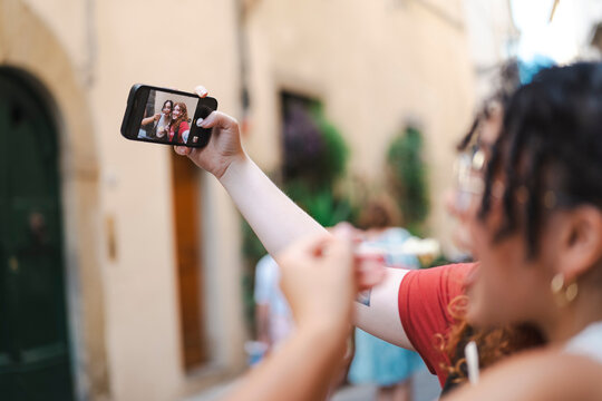 Two happy young female friends take a cheerful selfie on a smartphone while eating ice cream together on a scenic European street during a summer vacation trip.