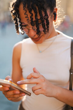 A young woman with curly hair and glasses focuses on her smartphone screen during golden hour. She is outdoors, typing with her finger and wearing wireless earbuds.