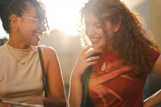 Two diverse young women with curly hair smile and laugh together during golden hour, sharing a warm moment of genuine friendship and happiness outdoors.
