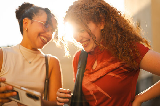 Two young diverse female friends laugh while looking at a smartphone screen together on a sunny city street. Sun flare highlights their joyful expressions.
