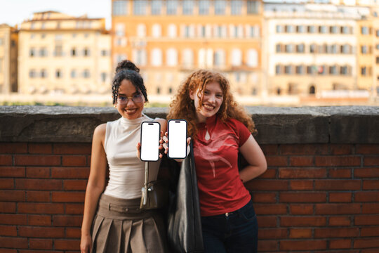 Two happy young female friends smile while holding up two smartphones with blank screens for a mockup, standing outdoors in front of a historic European city.