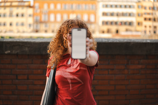 A young woman with curly red hair holds a smartphone with a blank mockup screen directly in front of her face, standing outdoors against a brick wall background.