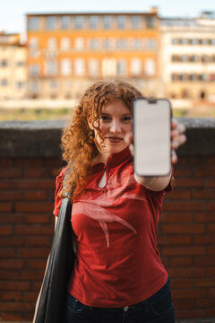 A smiling young woman with curly red hair holds up a mobile smartphone with a blank white screen towards the camera while standing outdoors in a European city.