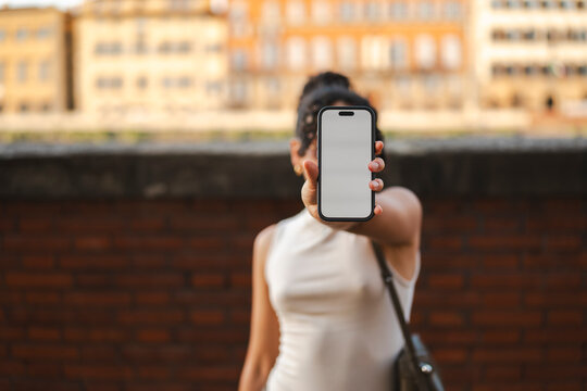 A woman stands outdoors in a city, holding up a smartphone with a blank white screen directly in front of her face. This screen serves as a clean digital mockup.