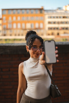Smiling young biracial woman with curly hair and glasses shows a smartphone with a blank screen for mockup space, posing outdoors with a historic city background.