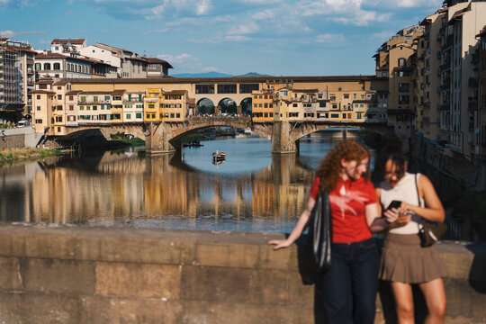 Two young female tourists use a smartphone on a bridge in Florence, Italy. The historic Ponte Vecchio bridge is beautifully reflected in the calm river water.