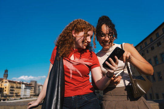 Two happy young female friends, one with curly red hair, laugh and smile while looking at a smartphone screen together outdoors on a sunny day in a historic city.