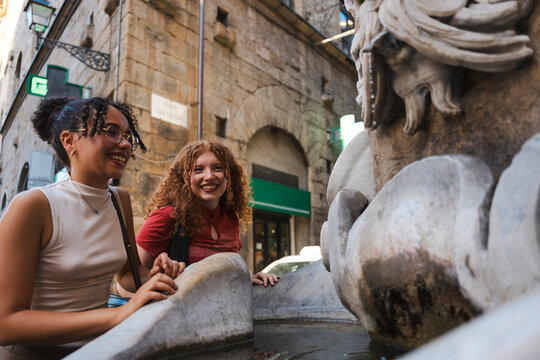 Two happy diverse young female friends smile while looking at an ancient stone water fountain as they explore a historic European city street together on vacation.