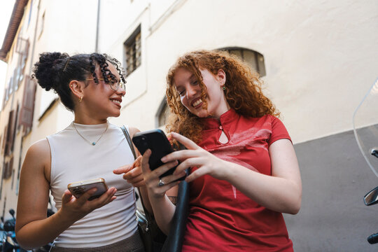 Two diverse young women with curly hair smiling and looking at a smartphone screen while standing in a city street. They are sharing a happy moment outdoors.