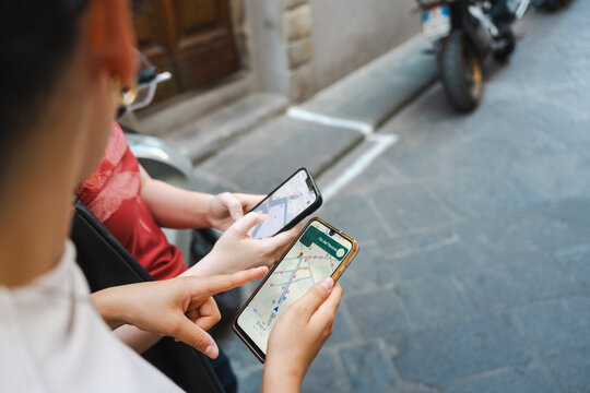 Two female travelers use navigation apps on their smartphones to find directions, pointing at the screens while walking down a historic European city street.