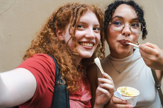 Two diverse young female friends with beautiful curly hair smile happily while taking a selfie portrait and eating sweet cold gelato from a cup on a sunny day.
