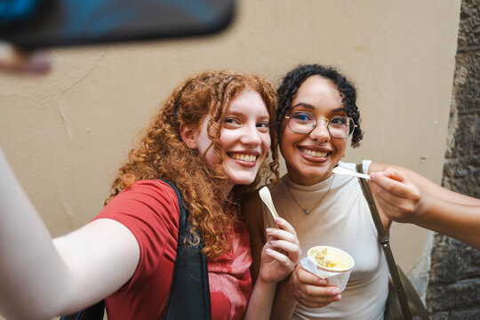 Two diverse, happy young female friends smile warmly while taking a selfie together. They are sharing a cup of delicious gelato outdoors on a sunny summer day.