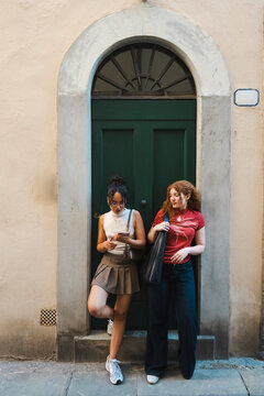 Two stylish young female friends stand together by an arched green wooden door in an ancient European street, checking a smartphone screen during their walk.