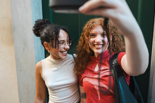 Two happy diverse female friends, one with curly black hair and glasses, and the other with curly red hair, smile and laugh while taking a selfie on a city street.