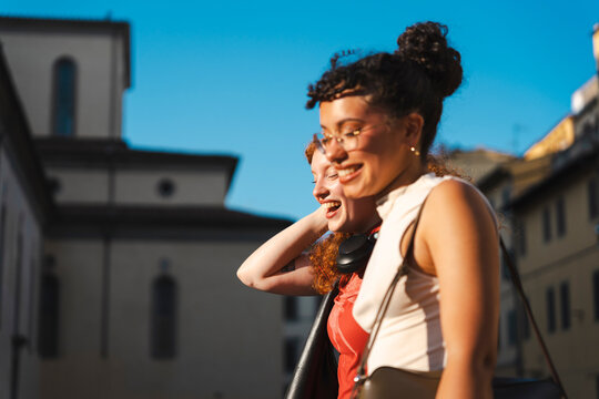 Two happy and stylish young female friends laugh together while walking outdoors in a historic European city on a sunny day, enjoying a moment of true friendship.