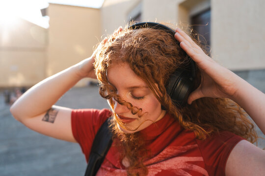 A young redhead woman with curly hair closes her eyes to enjoy listening to music on wireless headphones during a warm sunset golden hour outdoors in the city.