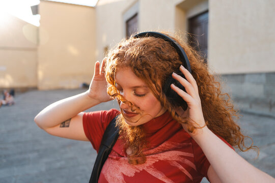 A cheerful young woman with long curly red hair closes her eyes and smiles, enjoying her favorite music on wireless headphones in a sunny city street at sunset.