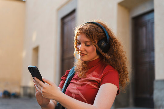 A smiling young woman with curly ginger hair wears black wireless headphones while looking at her mobile phone outdoors on a city street during golden hour.