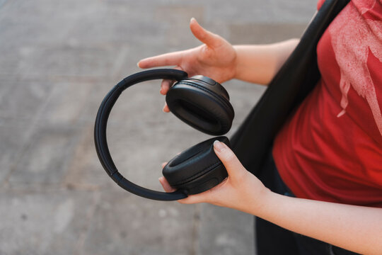 A close-up of a woman wearing a red t-shirt holding a pair of black wireless over-ear headphones in her hands while standing outside on a grey paved street.