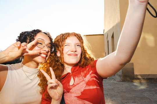 Two happy multiracial female friends with curly hair take a playful selfie outdoors. The cheerful young women smile and make peace signs on a sunny day.
