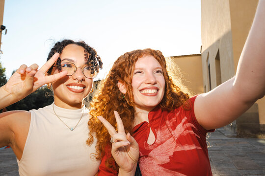 Two cheerful, diverse young women with curly hair smile happily while taking a selfie on a city street. They make peace signs in the warm golden hour sunlight.
