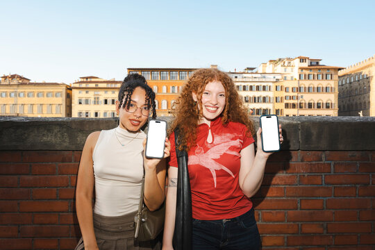 Two cheerful, diverse female friends stand outdoors on a sunny day. They smile as they show their smartphone screens to the camera against a classic city backdrop.