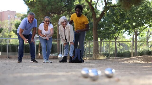  A group of multiracial middle aged mixed-sex adult people playing petanque game outdoors in public park on a sunny day