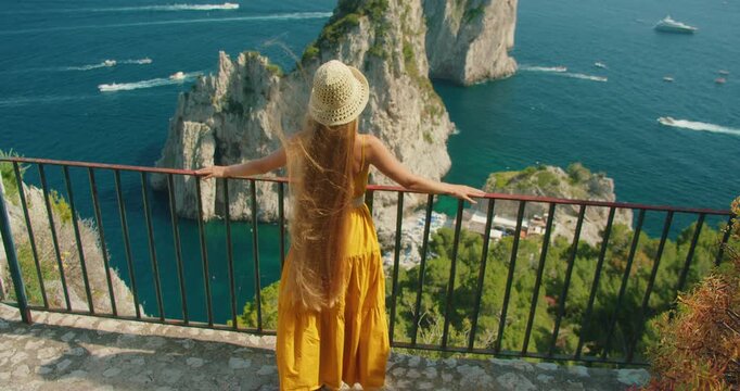 Back view of woman leaning on railing above Faraglioni cliffs and deep blue sea in Capri Italy