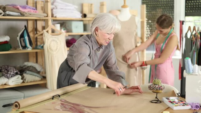 Attentive mature tailoress drawing dress design on paper while other working with mannequin in background. High quality 4k footage
