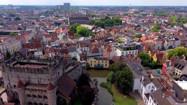 Aerial wide angle panoramic view of Gravensteen medieval stone castle keep and fortification walls overlooking a river canal park lawn and red tiled roofs under a summer sky