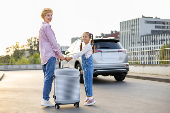A Caucasian mother and her daughter stand with a suitcase next to a car, ready for a trip