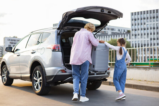 A Caucasian mother and her daughter are loading a suitcase into the trunk of a car, preparing for a journey