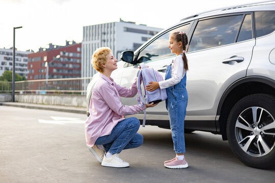 A mother hands a backpack to her daughter next to a car, preparing for school or an outing