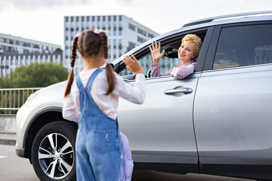 A smiling mother in a car waves goodbye to her daughter who is standing outside with a backpack