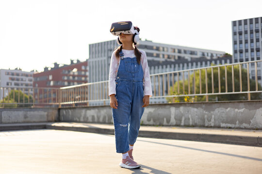 A young Caucasian girl wearing denim overalls and a VR headset explores a virtual world while standing on a rooftop with city buildings in the background