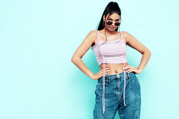 Young beautiful smiling female in trendy summer jeans and top clothes. Carefree woman posing near blue wall in studio. Sexy positive model having fun indoors. Cheerful and happy. In sunglasses © halayalex