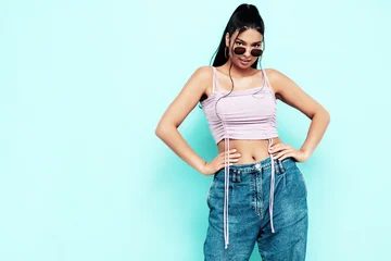 Young beautiful smiling female in trendy summer jeans and top clothes. Carefree woman posing near blue wall in studio. Sexy positive model having fun indoors. Cheerful and happy. In sunglasses © halayalex