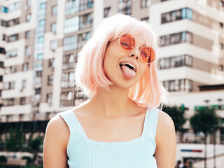 Fototapeta premium Young beautiful confident hipster female in trendy summer clothes. Sexy carefree woman posing in the street in wig. Positive funny model having at sunset. In sunglasses. At sunny day. Shows tongue