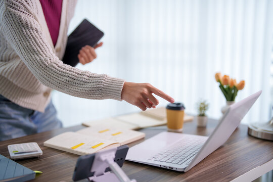 Adult woman reaching toward laptop while holding phone and notebook for online education studying at home workspace