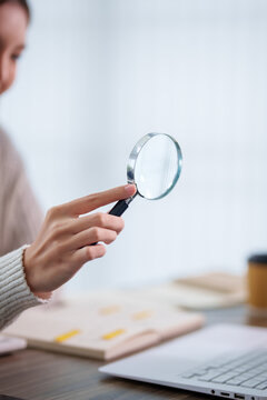 Adult woman holding magnifying glass examining documents at desk in bright office with copy space